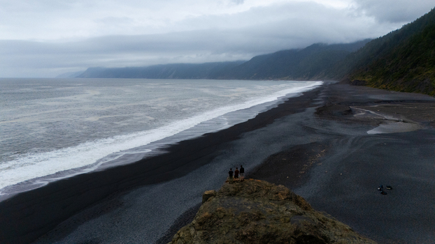 Aerial view of three people standing on a rocky outcrop overlooking the black sand beach and misty coastal mountains of the Lost Coast in Humboldt County California United States.