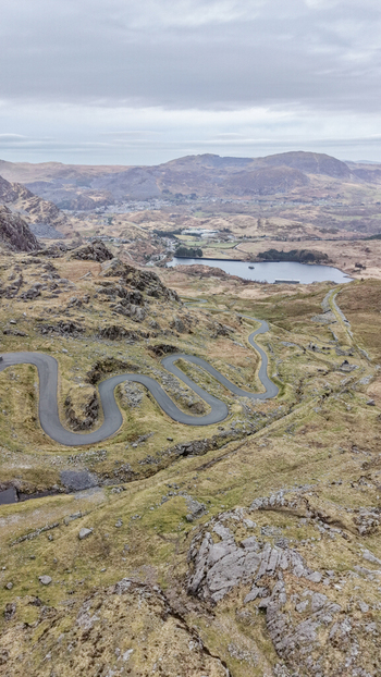 Aerial view of a winding road snaking through the rugged undulating terrain contrasting starkly with the serene lake nestled in the distance Blaenau Ffestiniog Wales United Kingdom. Aerial view of a winding road snaking through the rugged undulating terrain contrasting starkly with the serene lake nestled in the distance Blaenau Ffestiniog Wales United Kingdom.