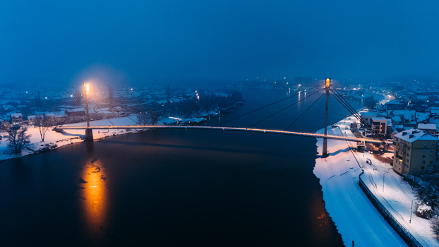 Aerial view of the illuminated St. Irenaeus Bridge spanning the Sava River at dusk with snow covered banks in Sremska Mitrovica Vojvodina Serbia.