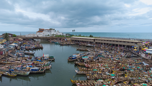Aerial view of Elmina Castle overlooking a bustling harbor filled with numerous colorful wooden fishing boats along the coast in Elmina Central Region Ghana. Aerial view of Elmina Castle overlooking a bustling harbor filled with numerous colorful wooden fishing boats along the coast in Elmina Central Region Ghana.