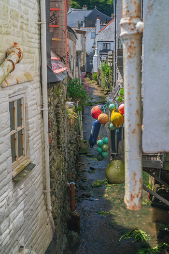 View of a narrow stream flowing between historic stone cottages featuring colorful fishing buoys hanging near the water in Polperro Cornwall United Kingdom. View of a narrow stream flowing between historic stone cottages featuring colorful fishing buoys hanging near the water in Polperro Cornwall United Kingdom.