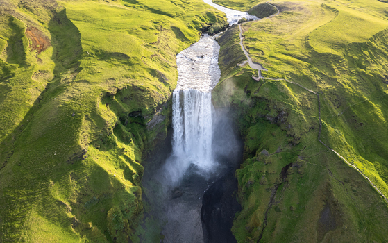 Aerial view of Skogafoss waterfall cascading down a steep cliff surrounded by lush green hills in Rangarthing eystra Iceland.