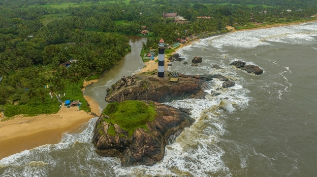 Aerial view of Kapu Light House standing on rocky outcrops surrounded by the Arabian Sea with lush palm trees and a river meeting the coast in Udupi Karnataka India.