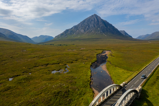 Aerial view of Buachaille Etive Mor mountain peak rising above the green landscape of Glen Coe with a winding river and road bridge in Ballachulish Scotland United Kingdom. Aerial view of Buachaille Etive Mor mountain peak rising above the green landscape of Glen Coe with a winding river and road bridge in Ballachulish Scotland United Kingdom.