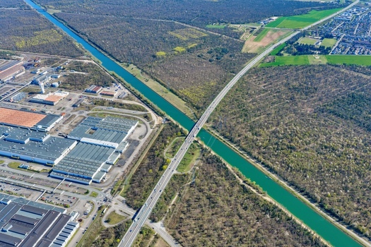 Aerial view of the stark turquoise canal slicing through verdant forests bisected by a gray concrete bridge near industrial buildings Sausheim Grand Est France.