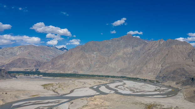 Aerial view of Shigar Valley featuring a braided river flowing through a dry riverbed surrounded by rugged mountains and green vegetation under a clear blue sky in Shigar Gilgit Pakistan.