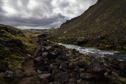 View of a river rushes through a rugged landscape of dark volcanic rocks and moss covered hills under a dramatic sky Skaftárhreppur Iceland. View of a river rushes through a rugged landscape of dark volcanic rocks and moss covered hills under a dramatic sky Skaftárhreppur Iceland.
