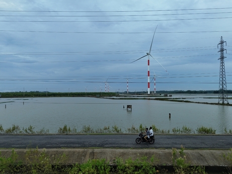 Aerial view of Khurushkul wind farm with turbines above flooded fields and a road with a motorcycle carrying two people under a cloudy sky in Cox s Bazar Chittagong Division Bangladesh. Aerial view of Khurushkul wind farm with turbines above flooded fields and a road with a motorcycle carrying two people under a cloudy sky in Cox s Bazar Chittagong Division Bangladesh.