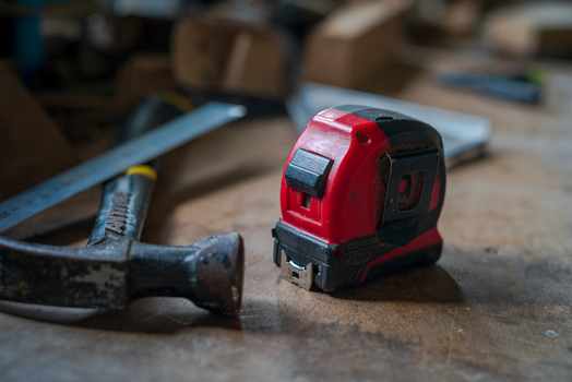 View of a red tape measure resting on a wooden workbench alongside a hammer and a metal ruler in Looe Cornwall United Kingdom. View of a red tape measure resting on a wooden workbench alongside a hammer and a metal ruler in Looe Cornwall United Kingdom.