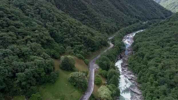 Aerial view of a winding road with a red car alongside a rushing river flowing through a lush green mountain valley with dense forest. Val Verzasca Switzerland. Aerial view of a winding road with a red car alongside a rushing river flowing through a lush green mountain valley with dense forest. Val Verzasca Switzerland.