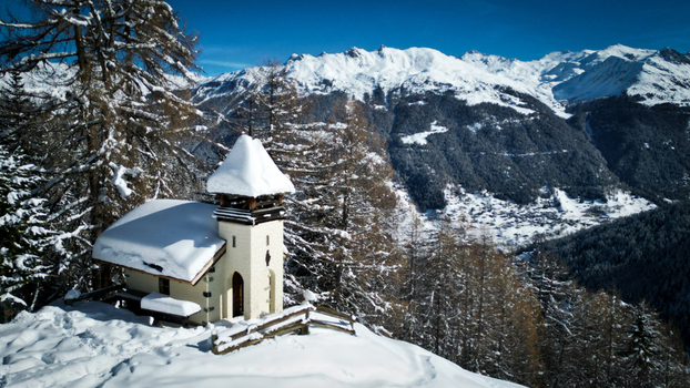 Aerial view of a snow laden chapel stands serenely against a backdrop of majestic snow capped mountains Grimentz Valais Switzerland.