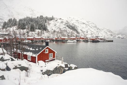 View of traditional red wooden rorbu cabins situated on the rocky shoreline of a fjord surrounded by snow covered mountains and pine trees in Leknes Lofoten Norway.