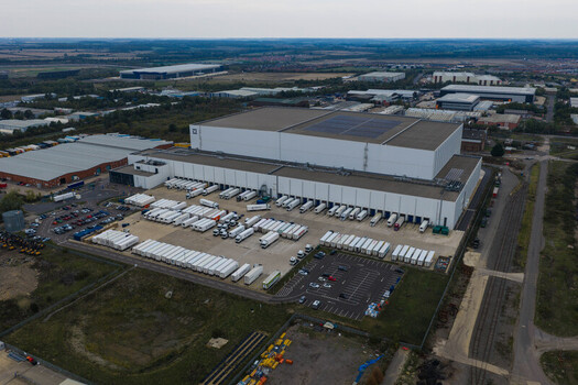 Aerial view of a large logistics warehouse facility with numerous semi trailers parked at loading bays and in the parking lot in an industrial area in the United Kingdom. Aerial view of a large logistics warehouse facility with numerous semi trailers parked at loading bays and in the parking lot in an industrial area in the United Kingdom.