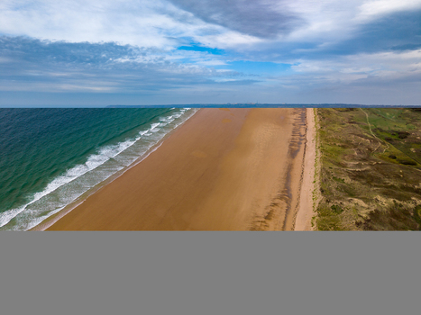 Aerial view of the Dunes de Biville featuring a vast sandy beach rolling ocean waves and grassy coastal dunes under a cloudy sky in La Hague Normandie France.
