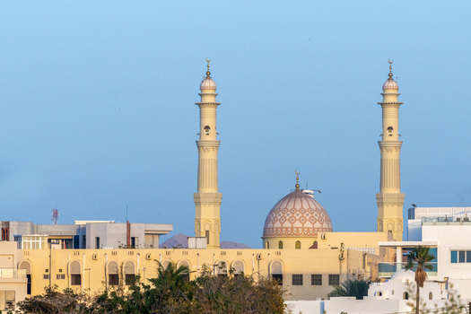 View of a mosque with two tall minarets and a patterned dome rising above urban buildings in Muscat Muscat Governorate Oman. View of a mosque with two tall minarets and a patterned dome rising above urban buildings in Muscat Muscat Governorate Oman.