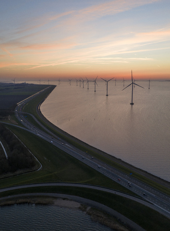 Aerial view of offshore wind turbines in the IJsselmeer lake standing next to a coastal highway dike during a colorful sunset Netherlands.