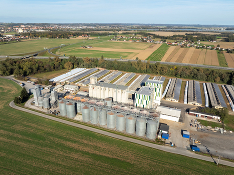 Aerial view of a large industrial agricultural facility with grain silos and long warehouses surrounded by green fields under a clear blue sky in Slovenia.