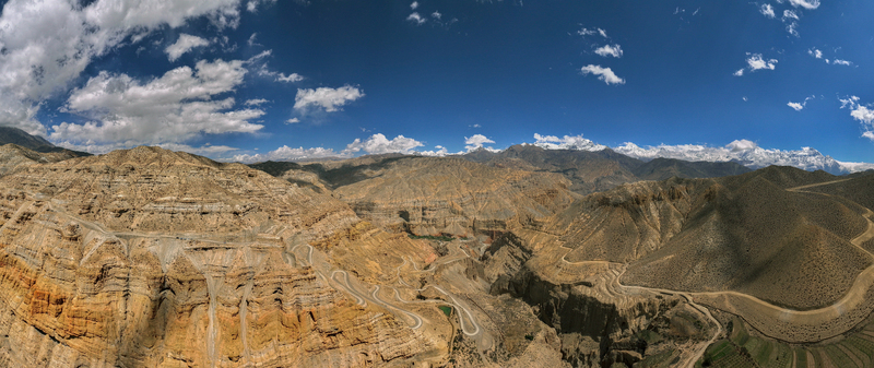 Aerial view of a winding road traversing the arid rugged mountain terrain of the Himalayas under a bright blue sky with scattered clouds in Nepal.