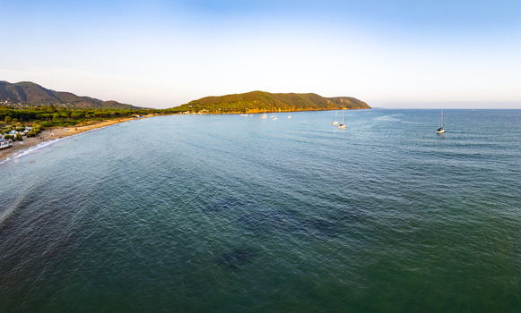 Aerial view of sailboats anchored in the calm turquoise waters of Lacona Bay with a sandy beach and rolling hills in the background in Lacona Tuscany Italy.