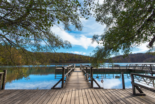 View of a wooden pier leading into the calm waters of Plitvice Lakes National Park surrounded by autumn forest and trees in Croatia.
