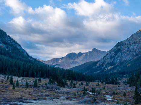 Aerial view of a mountain valley featuring dense pine forests a winding stream and rugged peaks under a dramatic cloudy sky in Colorado United States. Aerial view of a mountain valley featuring dense pine forests a winding stream and rugged peaks under a dramatic cloudy sky in Colorado United States.