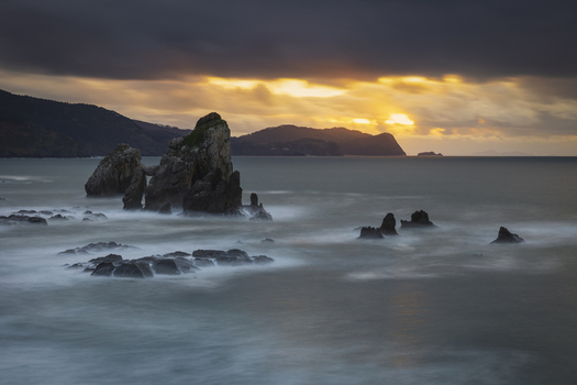 View of dramatic rock formations in the sea with misty waves under a golden sunset sky Gaztelugatxeko Doniene Euskadi Spain. View of dramatic rock formations in the sea with misty waves under a golden sunset sky Gaztelugatxeko Doniene Euskadi Spain.