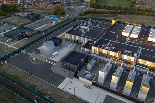 Aerial view of a modern data center facility featuring rooftop cooling units industrial equipment and perimeter fencing in the United Kingdom. Aerial view of a modern data center facility featuring rooftop cooling units industrial equipment and perimeter fencing in the United Kingdom.