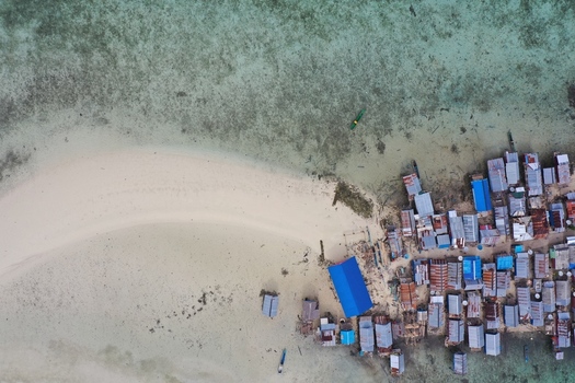 Aerial view of Pulau Maginti featuring traditional stilt houses built over shallow turquoise water along a white sand beach in Maginti South East Sulawesi Indonesia.