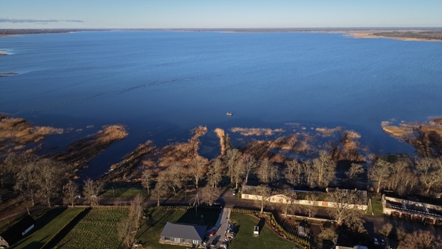 Aerial view of Lake Burtnieks with its calm blue waters surrounding reed beds bare trees and nearby rural buildings in Burtnieki Valmiera Municipality Latvia.