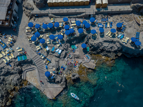 Aerial view of a rocky beach club with blue umbrellas sun loungers and people swimming in crystal clear turquoise water in Capri Campania Italy.