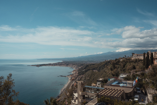 View of the azure sea meeting the rugged coastline under a vast sky with buildings nestled among lush greenery Taormina Catania Sicily Italy. View of the azure sea meeting the rugged coastline under a vast sky with buildings nestled among lush greenery Taormina Catania Sicily Italy.