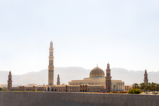 View of the Sultan Qaboos Grand Mosque with its prominent minaret and large dome set against a backdrop of mountains in Muscat Muscat Governorate Oman. View of the Sultan Qaboos Grand Mosque with its prominent minaret and large dome set against a backdrop of mountains in Muscat Muscat Governorate Oman.