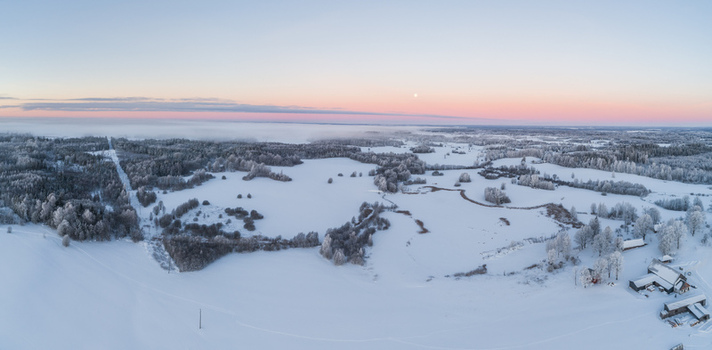 Aerial view of a vast snow covered landscape featuring frozen fields dense frost covered forests and a small farmhouse under a soft winter sky in Latvia. Aerial view of a vast snow covered landscape featuring frozen fields dense frost covered forests and a small farmhouse under a soft winter sky in Latvia.