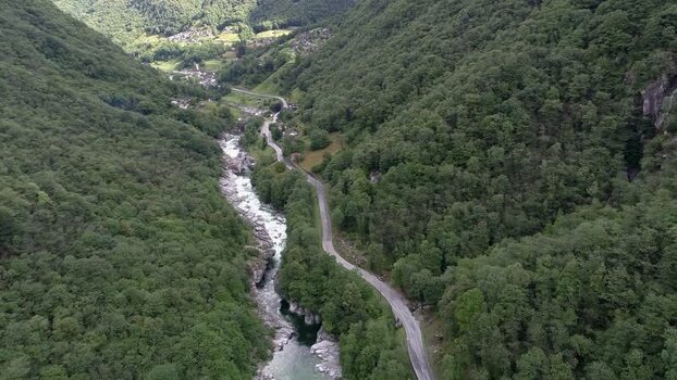 Aerial view of a winding road and a rocky river flowing through the lush green Verzasca Valley with dense forests and steep mountain slopes. Aerial view of a winding road and a rocky river flowing through the lush green Verzasca Valley with dense forests and steep mountain slopes.