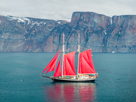 Aerial view of a sailboat with vibrant red sails navigating the deep blue waters of a bay set against the dramatic rocky cliffs of Uummannaq Kitaa Greenland. Aerial view of a sailboat with vibrant red sails navigating the deep blue waters of a bay set against the dramatic rocky cliffs of Uummannaq Kitaa Greenland.
