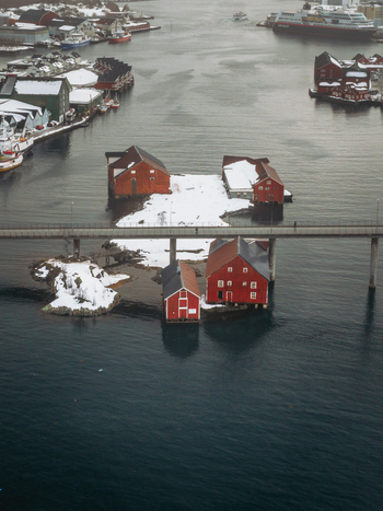 Aerial view of vibrant red cabins nestled on snowy islets linked by a slender bridge over the dark waters contrasting against the distant town Svolv&aelig;r Nordland Norway.