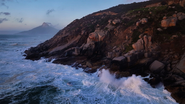 Aerial view of crashing waves against a rugged rocky coastline and steep cliffs with a mountain peak in the distance under a twilight sky Cape Town Western Cape South Africa. Aerial view of crashing waves against a rugged rocky coastline and steep cliffs with a mountain peak in the distance under a twilight sky Cape Town Western Cape South Africa.
