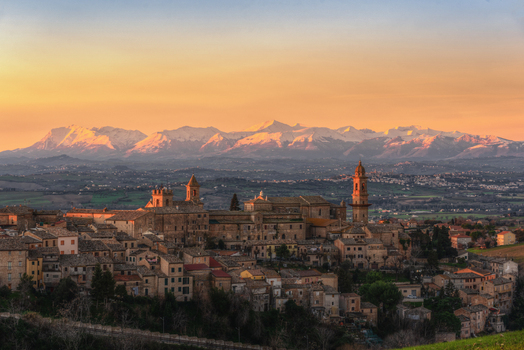 View of historic stone buildings and a church tower set against a backdrop of snow capped mountains under a warm sunset sky in Morrovalle Marche Italy.