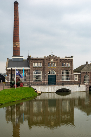 View of the Dutch Steam Engine Museum with its tall brick chimney and historic facade reflected in the canal water in Medemblik Noord Holland Netherlands. View of the Dutch Steam Engine Museum with its tall brick chimney and historic facade reflected in the canal water in Medemblik Noord Holland Netherlands.