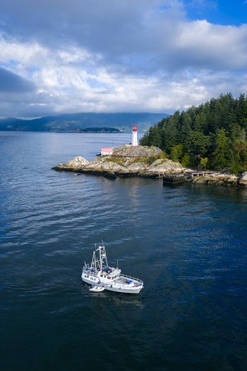 Aerial view of Point Atkinson Lighthouse on a rocky headland with a fishing boat in the foreground surrounded by evergreen trees and water West Vancouver British Columbia Canada.