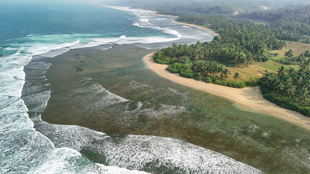 Aerial view of a tropical beach with turquoise shallow water coral reef crashing waves and lush palm trees along the coastline in Indonesia. Aerial view of a tropical beach with turquoise shallow water coral reef crashing waves and lush palm trees along the coastline in Indonesia.