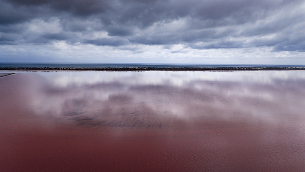 Aerial view of a vibrant pink salt lake with dramatic dark clouds reflecting on the water surface and a distant coastline in Australia. Aerial view of a vibrant pink salt lake with dramatic dark clouds reflecting on the water surface and a distant coastline in Australia.