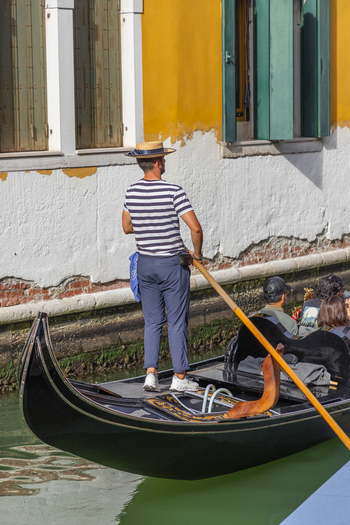 Venice Italy 21 September 2026 View of a gondolier in a striped shirt and straw hat steering a gondola along a canal with passengers and a yellow building in the background. Venice Italy 21 September 2026 View of a gondolier in a striped shirt and straw hat steering a gondola along a canal with passengers and a yellow building in the background.
