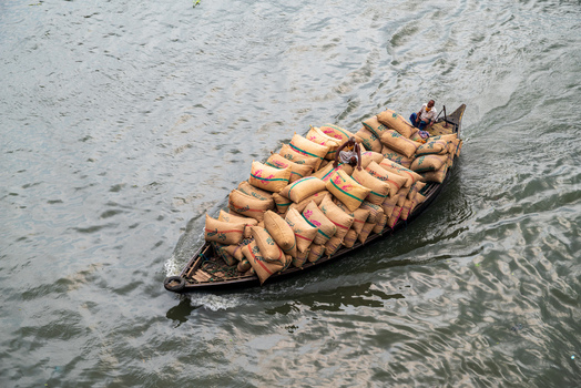 Dhaka Division Bangladesh 14 May 2026 Aerial view of a wooden boat heavily loaded with jute sacks and two men navigating the Buriganga River.