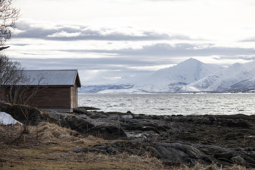 View of a traditional wooden boathouse on a rocky shoreline overlooking a fjord and snow capped mountains in Tromso Troms Norway.