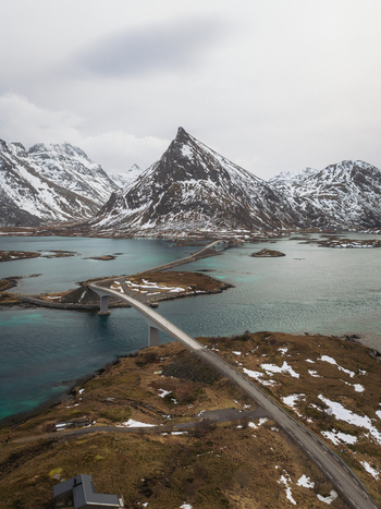 Aerial view of Flakstadbruene bridge stretches across the turquoise waters under the gaze of snow dusted peaks Flakstadbruene Nordland Norway. Aerial view of Flakstadbruene bridge stretches across the turquoise waters under the gaze of snow dusted peaks Flakstadbruene Nordland Norway.