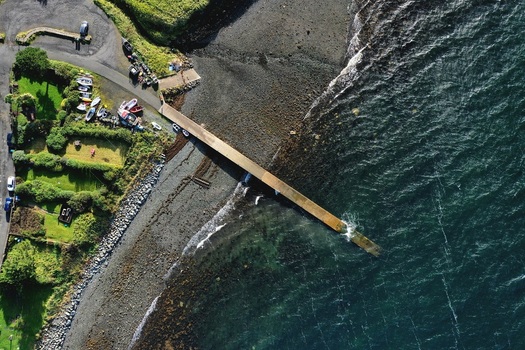 Aerial view of a concrete pier extending into the dark blue sea from a rocky beach with small boats and green coastal gardens in Stein Scotland United Kingdom. Aerial view of a concrete pier extending into the dark blue sea from a rocky beach with small boats and green coastal gardens in Stein Scotland United Kingdom.