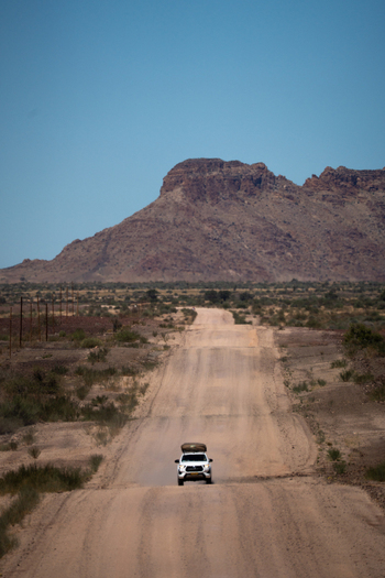 View of a lone white vehicle kicking up dust on a straight dirt road towards a looming sun baked mountain range Karas Region Keetmanshoop Namibia. View of a lone white vehicle kicking up dust on a straight dirt road towards a looming sun baked mountain range Karas Region Keetmanshoop Namibia.