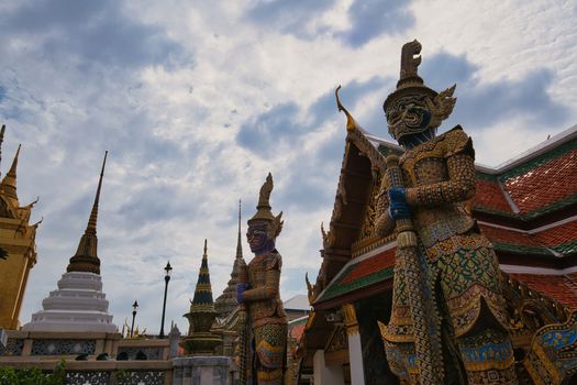View of The Grand Palace featuring ornate demon guardian statues traditional Thai architecture with golden spires and a cloudy sky Bangkok Thailand. View of The Grand Palace featuring ornate demon guardian statues traditional Thai architecture with golden spires and a cloudy sky Bangkok Thailand.