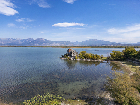 Aerial view of a building on a small peninsula in Lake Shkoder surrounded by calm water lush trees and distant mountains in Shiroka Shkoder County Albania. Aerial view of a building on a small peninsula in Lake Shkoder surrounded by calm water lush trees and distant mountains in Shiroka Shkoder County Albania.